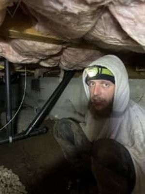 Person crouches in a dim crawlspace under exposed insulation, wearing a hood and headlamp.