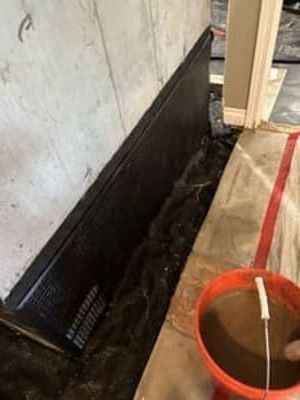 Basement wall with black waterproofing membrane, gravel trench, and orange bucket of muddy water