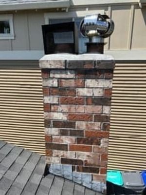 Brick chimney with a metal vent cap on a shingled roof beside a house wall.
