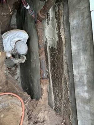 Worker inspecting a narrow trench beside a concrete wall, with exposed pipes and dirt debris