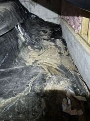 Black plastic sheeting covering insulation in a crawl space, with debris and a small opening at right.