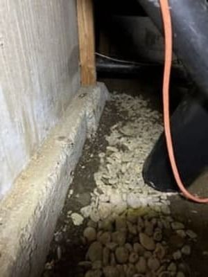 Narrow crawlspace with gravel on the floor, concrete wall, and an orange hose or cable hanging down
