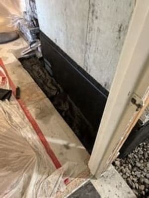 Interior wall with a long exposed trench at floor level beside a doorway, covered with protective plastic and tape.