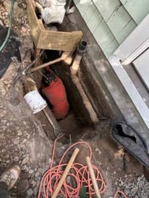Open utility trench beside a house with a red tank and coiled orange cable on the ground