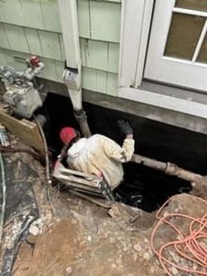 Worker in a trench beside a house, handling pipes and tools near the foundation.