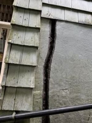 Cracked exterior wall beside green siding, with a black pipe crossing the bottom.