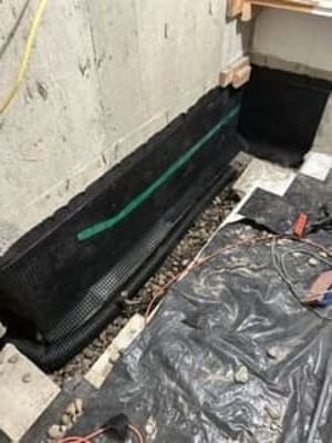 Construction area with black waterproofing along a wall and plastic sheeting on the floor.