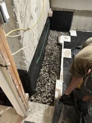 Worker installing black gravel drainage bed beside a house foundation with white edging blocks