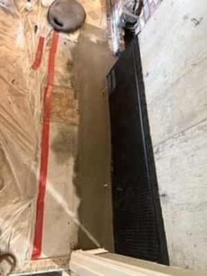 Narrow construction trench with a long black drain grate beside a concrete wall and marked floor lines