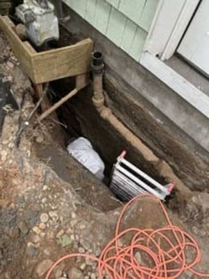 Open trench beside a house with exposed pipes, a wooden planter, and orange extension cords.