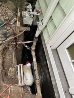 Narrow utility trench beside a house with exposed pipes, valves, and a large white tank.