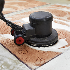 A floor buffing machine scrubs a brown, patterned carpet with white cleaning foam.