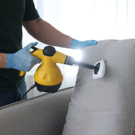 A person in gloves uses a yellow handheld steam cleaner to sanitize a gray fabric couch.