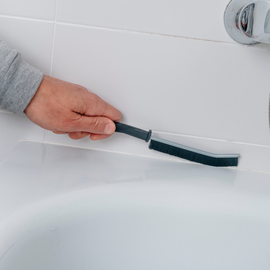 A person cleans white bathroom tiles and a bathtub edge with a long, thin, dark-bristled brush.