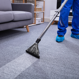A professional cleaner in blue uniform and shoe covers steam cleans a gray carpet in a living room.