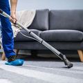 A worker in blue uniform and shoe covers uses a carpet cleaning wand to clean a gray carpet in front of a sofa.