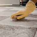 A hand wearing a yellow rubber glove uses a wooden scrub brush to clean gray textured floor tiles.