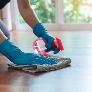 Hands wearing blue rubber gloves cleaning a wooden floor with a microfiber cloth and a red spray bottle.