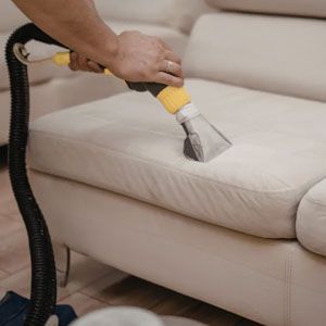 A hand uses an extraction vacuum tool to deep clean a light-colored fabric sofa.
