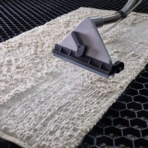 A white rug being cleaned by a professional steam cleaning nozzle on a black, honeycomb-patterned grid surface.