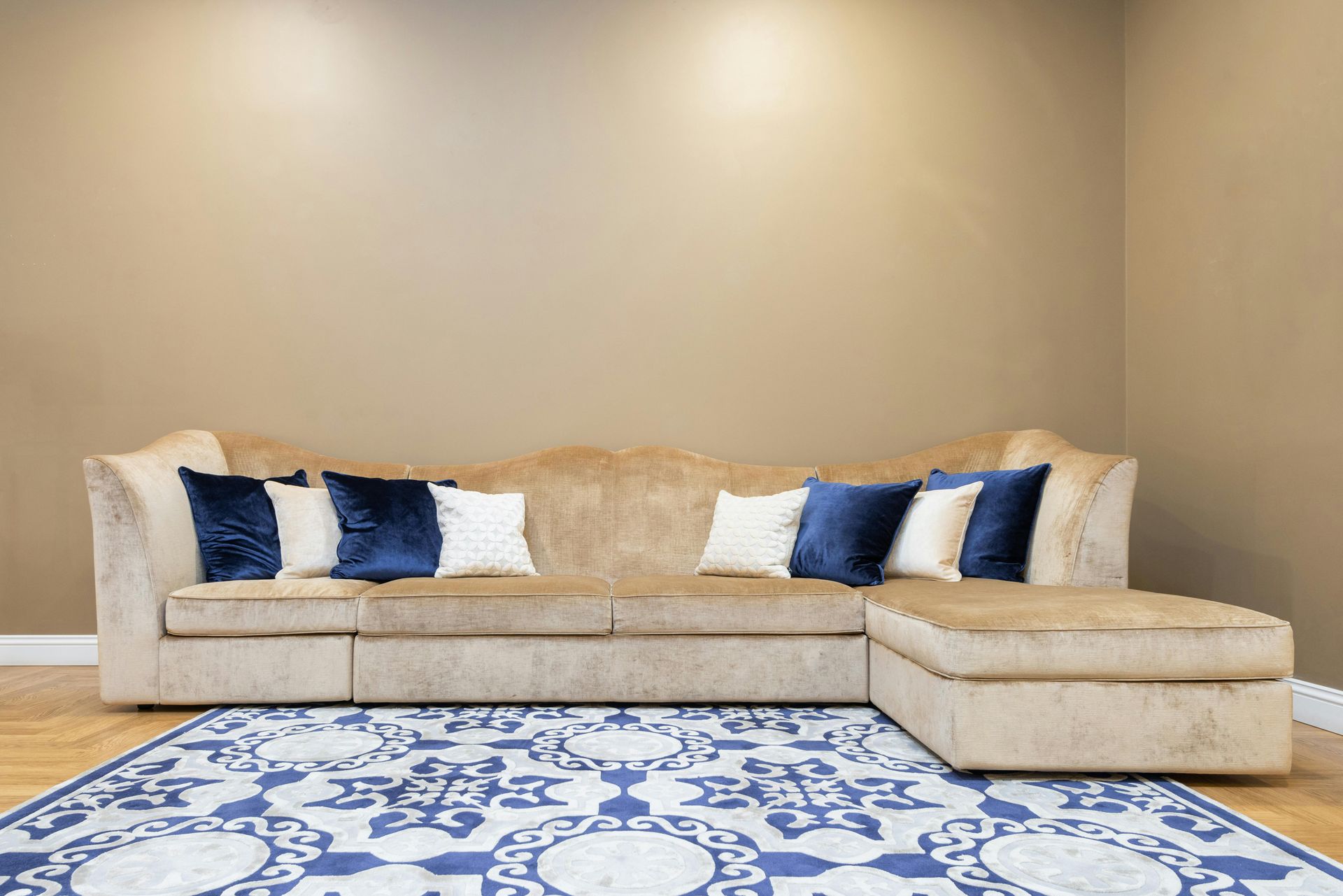 A beige sectional sofa with blue and white throw pillows sits on a blue and white patterned rug against a plain wall.