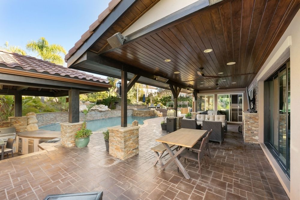 Covered patio with brick floor, wooden ceiling, stone columns, and outdoor dining tables overlooking a garden