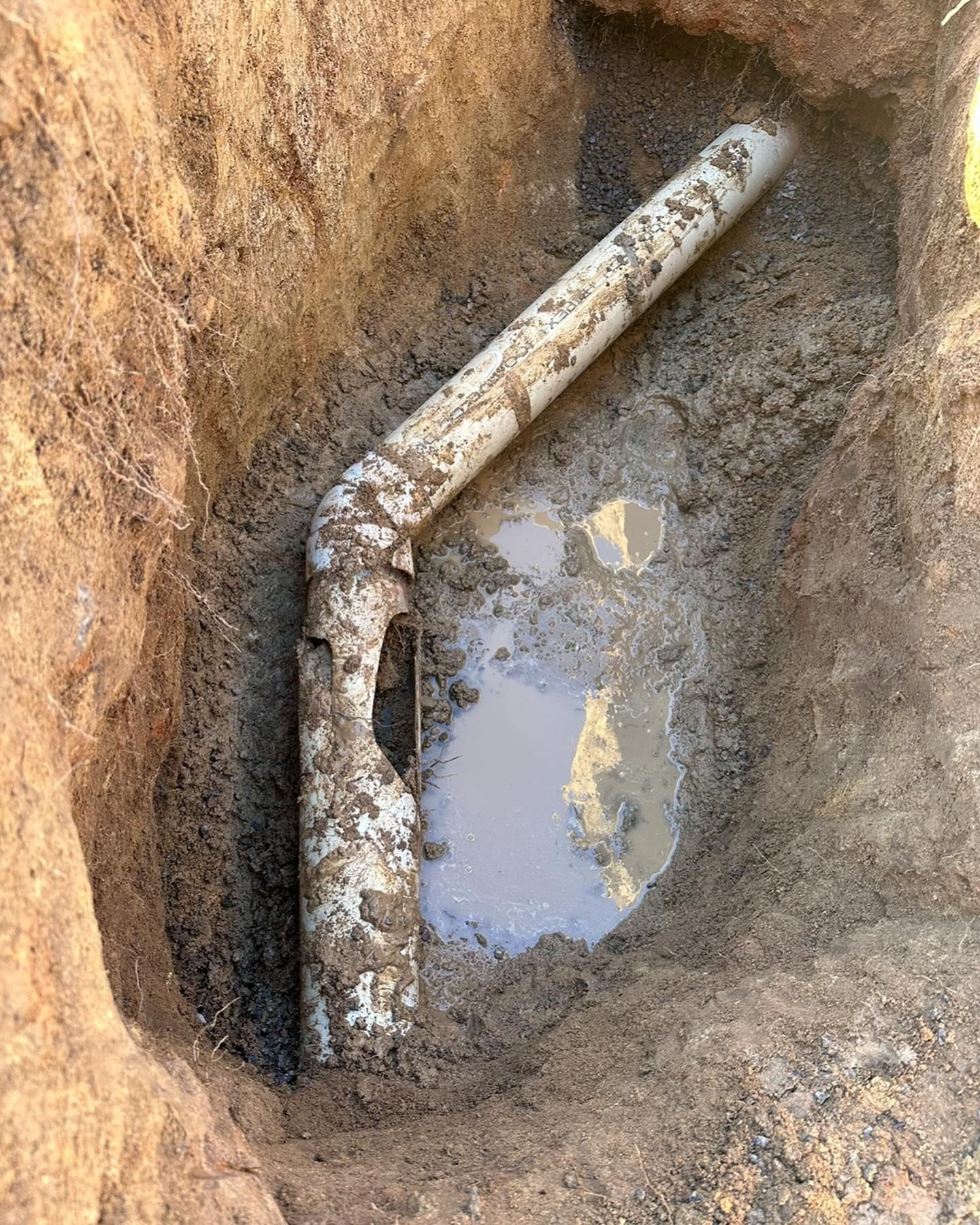 A broken PVC pipe angled in a dirt trench, surrounded by mud and a puddle of water — B3 Plumbing in Flynn, ACT