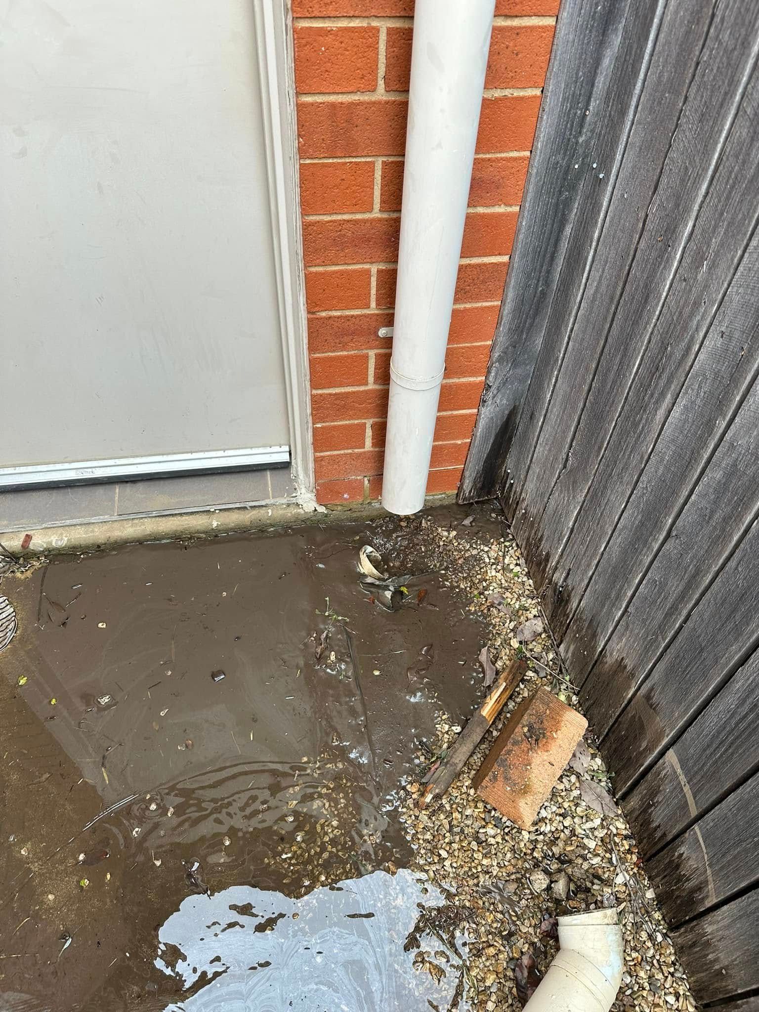 Wet patio corner with a downspout, brick wall, wooden fence, and scattered debris. Water pools on the surface — B3 Plumbing in Flynn, ACT