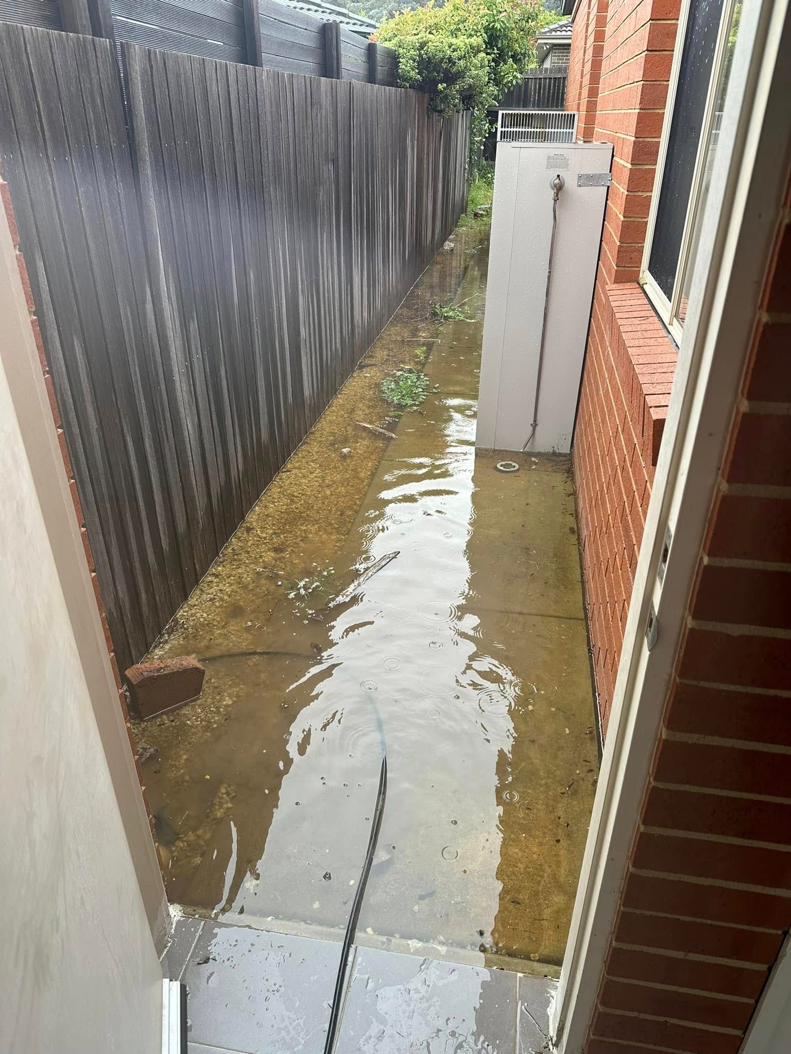 Flooded narrow outdoor space between a wooden fence and brick wall. Water covers the ground — B3 Plumbing in Flynn, ACT