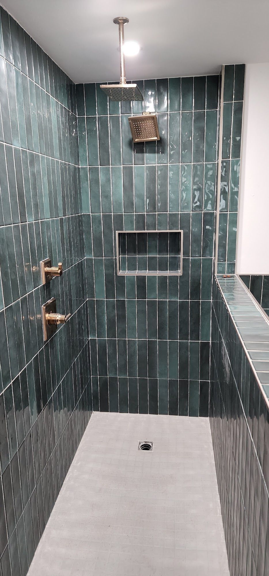 A modern shower with dark green tiles.  There is a shower head, faucet, and small built-in shelf.