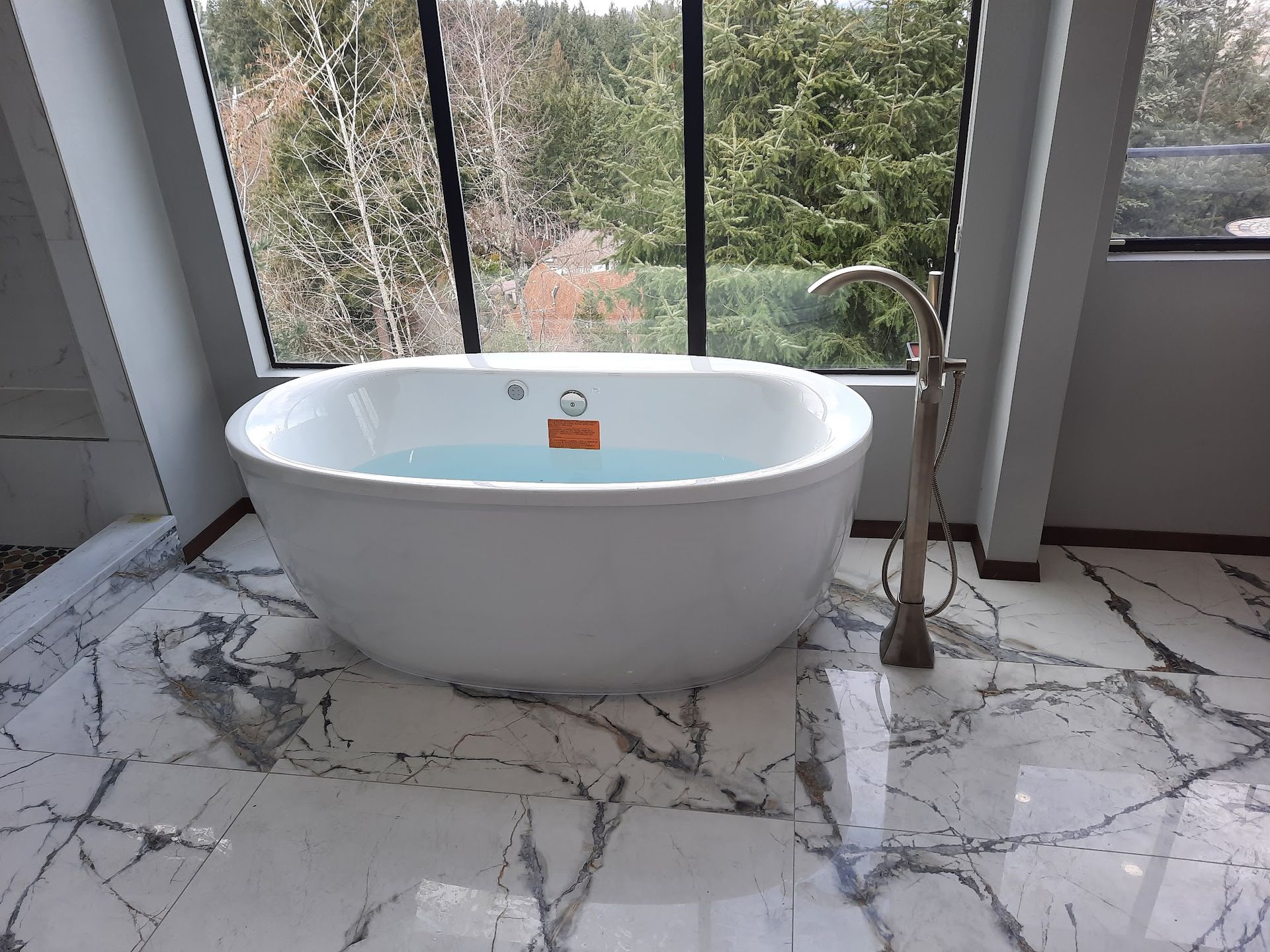 Oval white bathtub filled with water, next to a tall faucet, in a bathroom with large window overlooking trees.