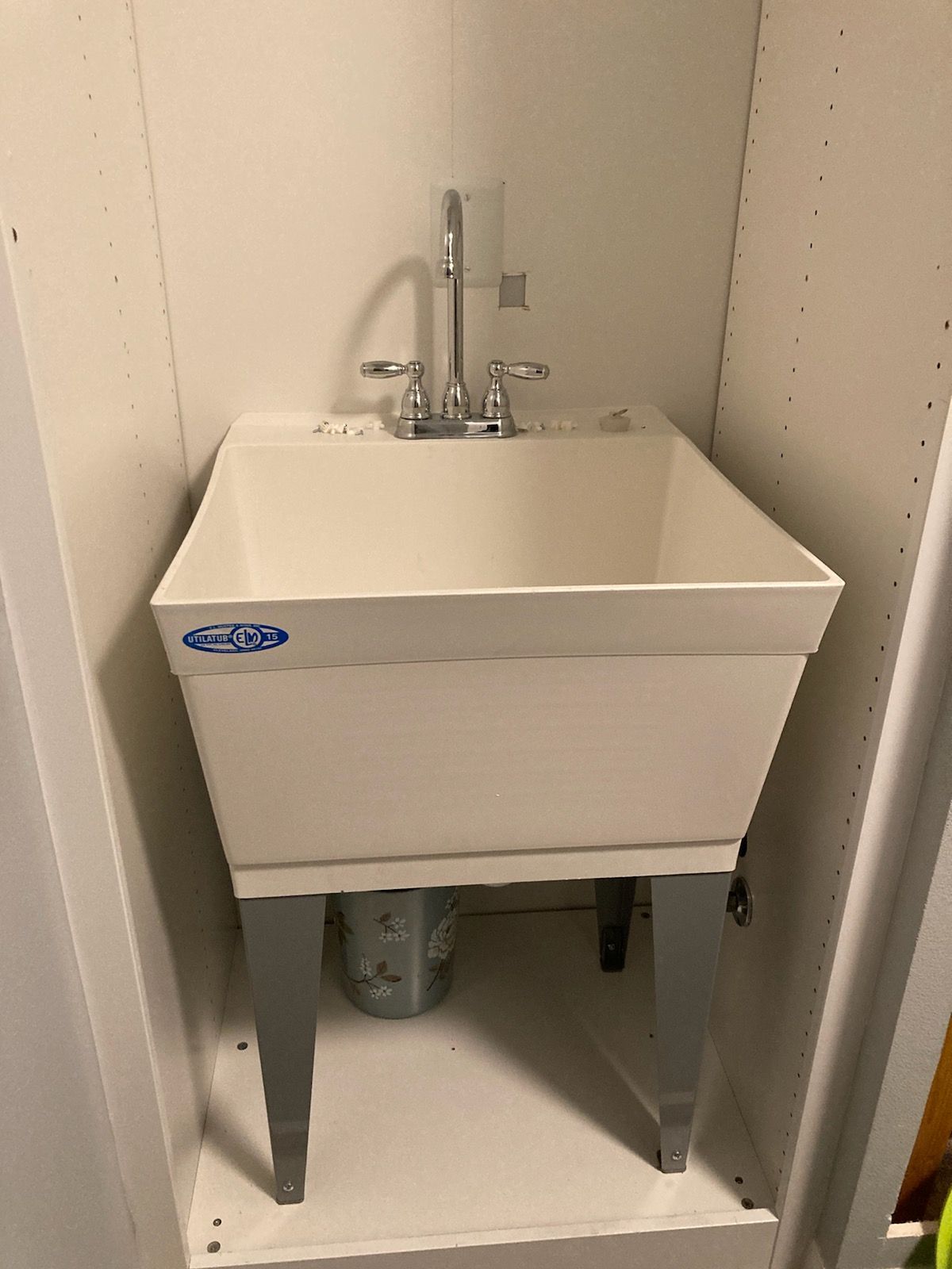 White utility sink with chrome faucet and legs, in a white-walled alcove.