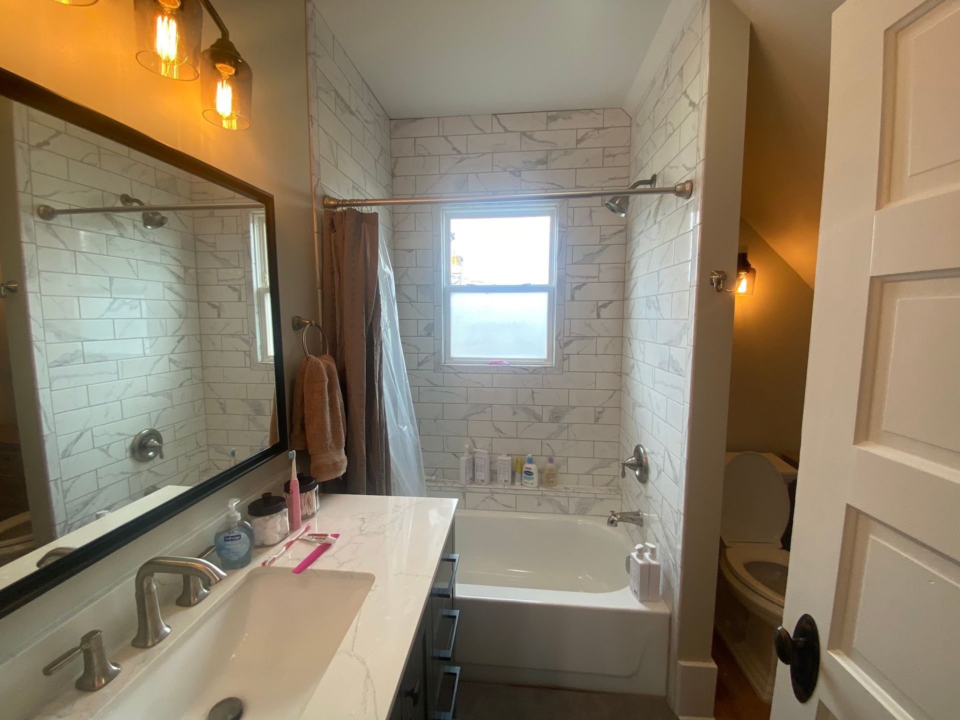 Bathroom with white tile shower, vanity with sink, toilet, and lit wall sconces.