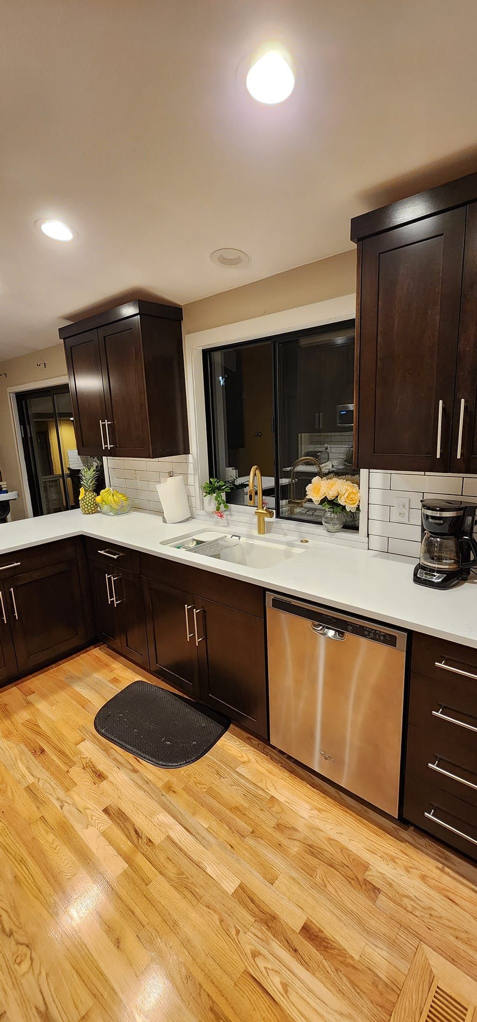 A kitchen with dark wood cabinets, light countertops, a stainless steel dishwasher, and hardwood floors.