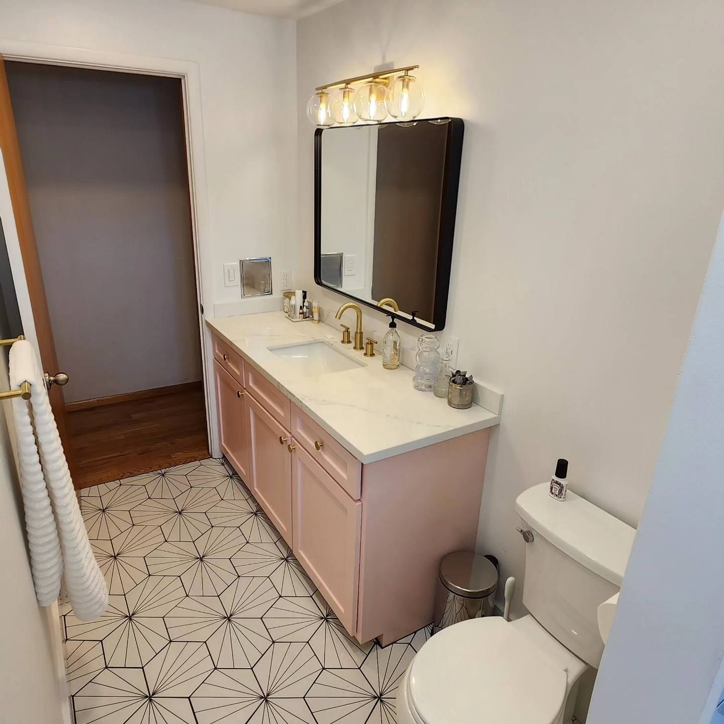 Bathroom with pink vanity, black-framed mirror, patterned floor tiles, and gold fixtures.