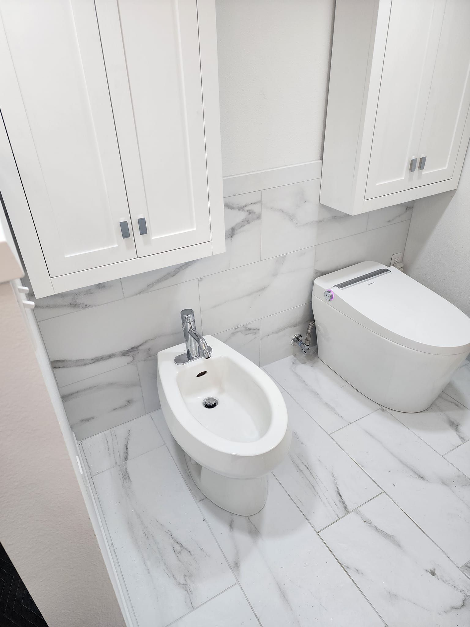 White bathroom with a bidet and a smart toilet under white cabinets. Marble-look tile.