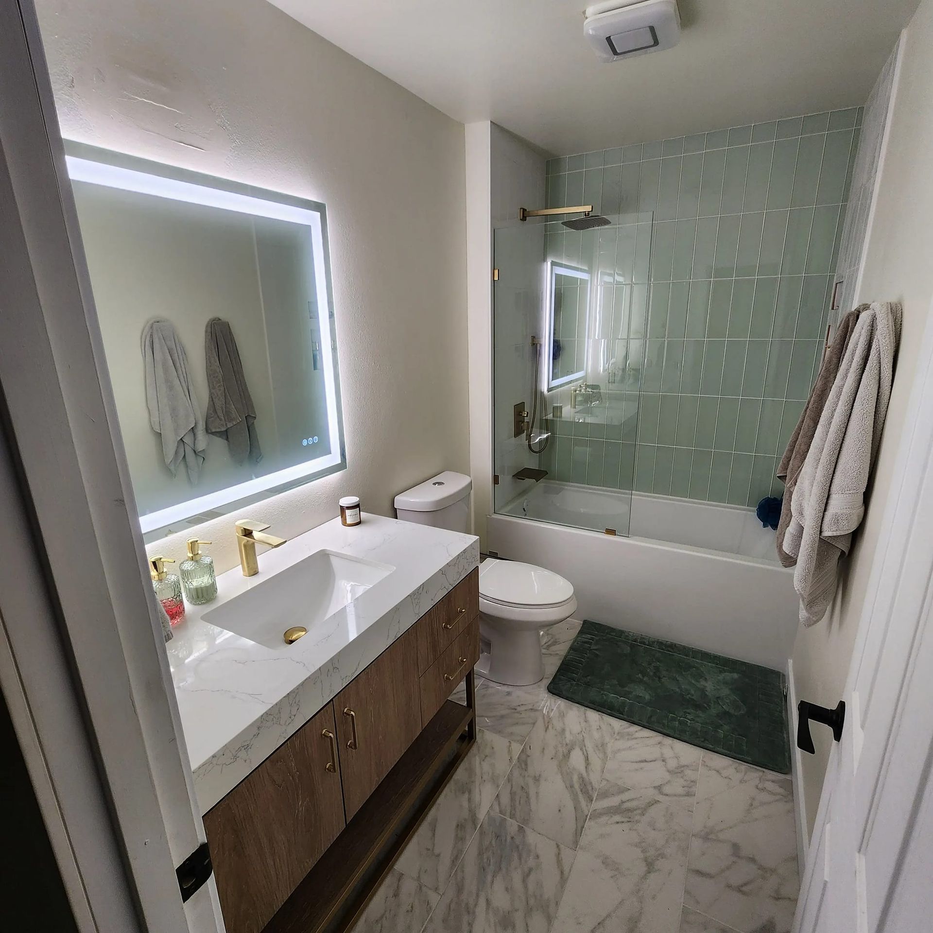 Bathroom with marble floors, gold fixtures, and a green tiled shower. A large, lit mirror hangs over a wood vanity.