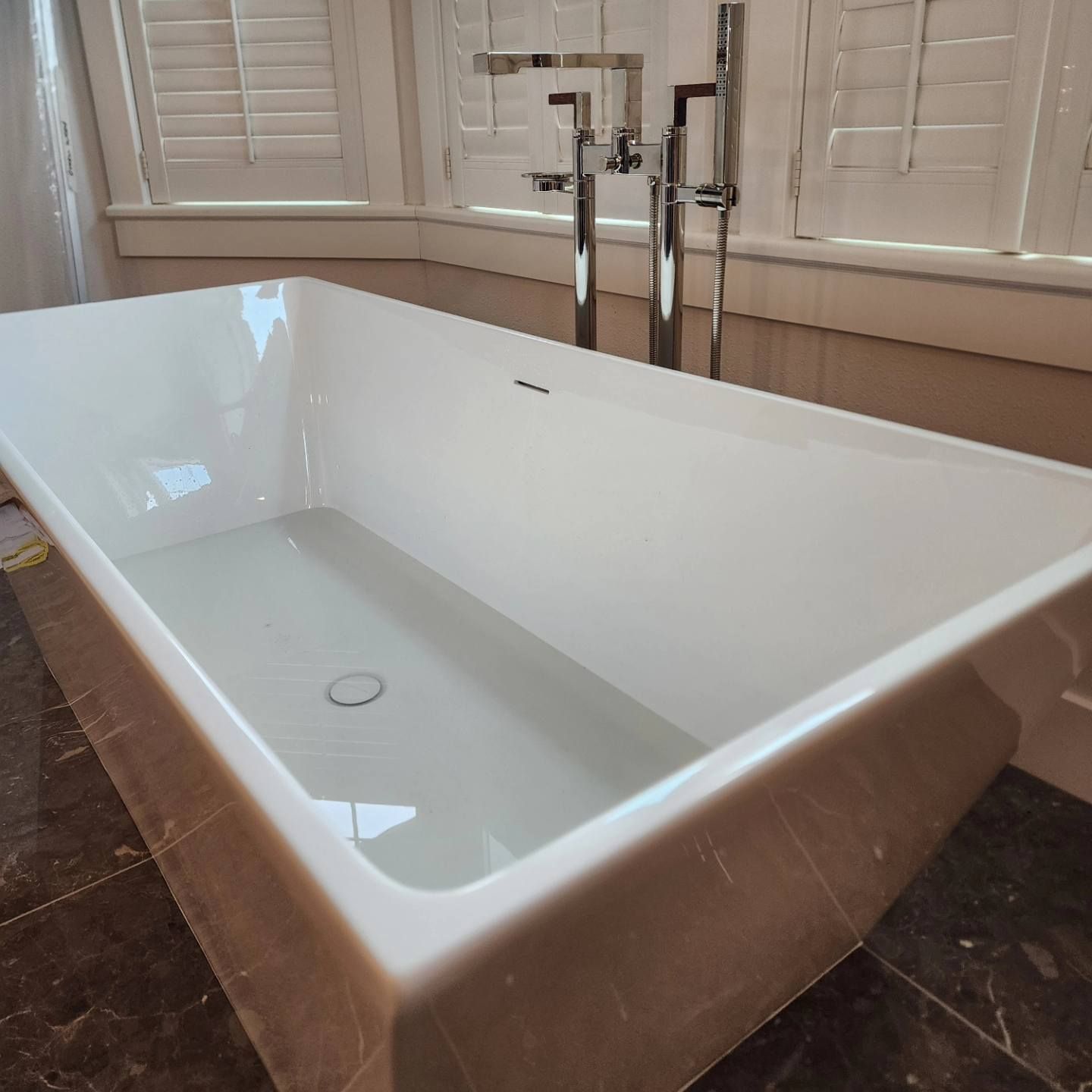 A rectangular white bathtub filled with water, chrome faucet, window with white blinds, brown tiled floor.