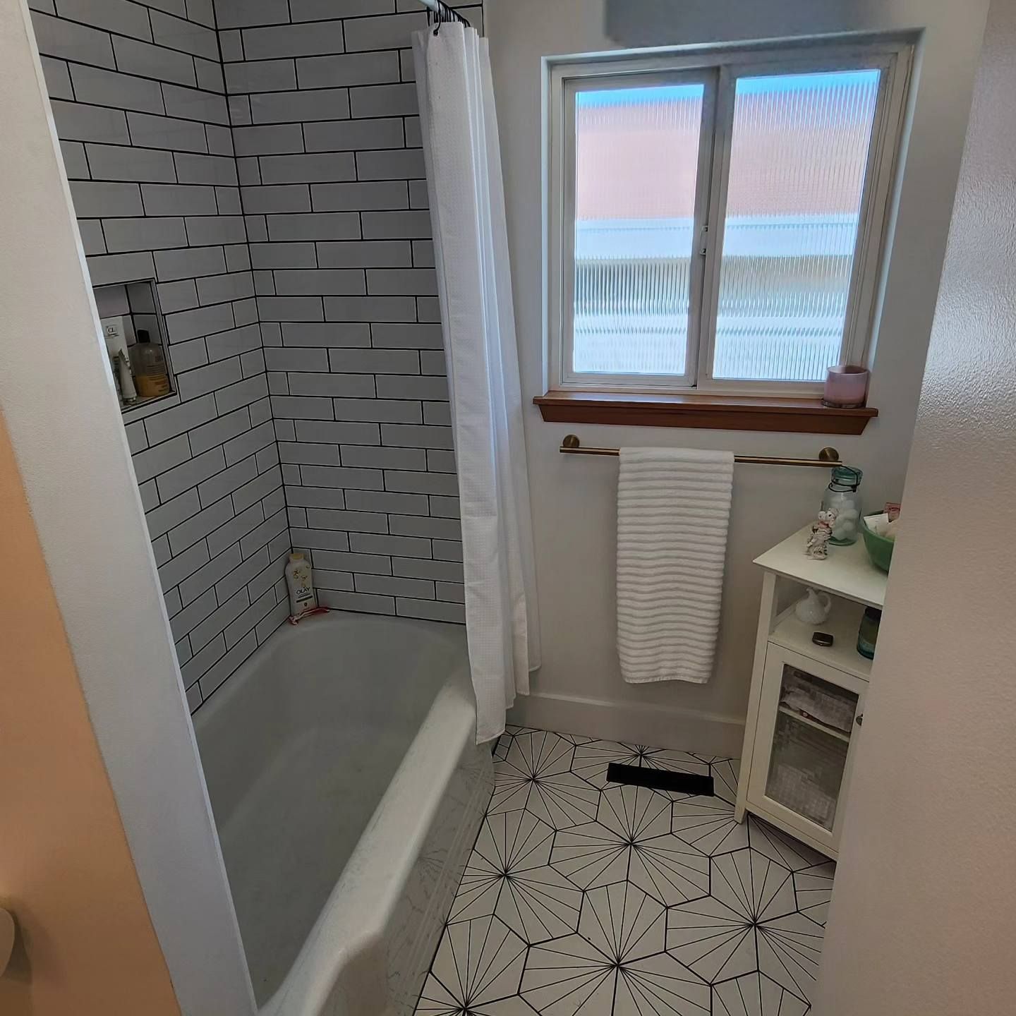 Bathroom with white tiled shower, window, towel rack, cabinet, and patterned floor.