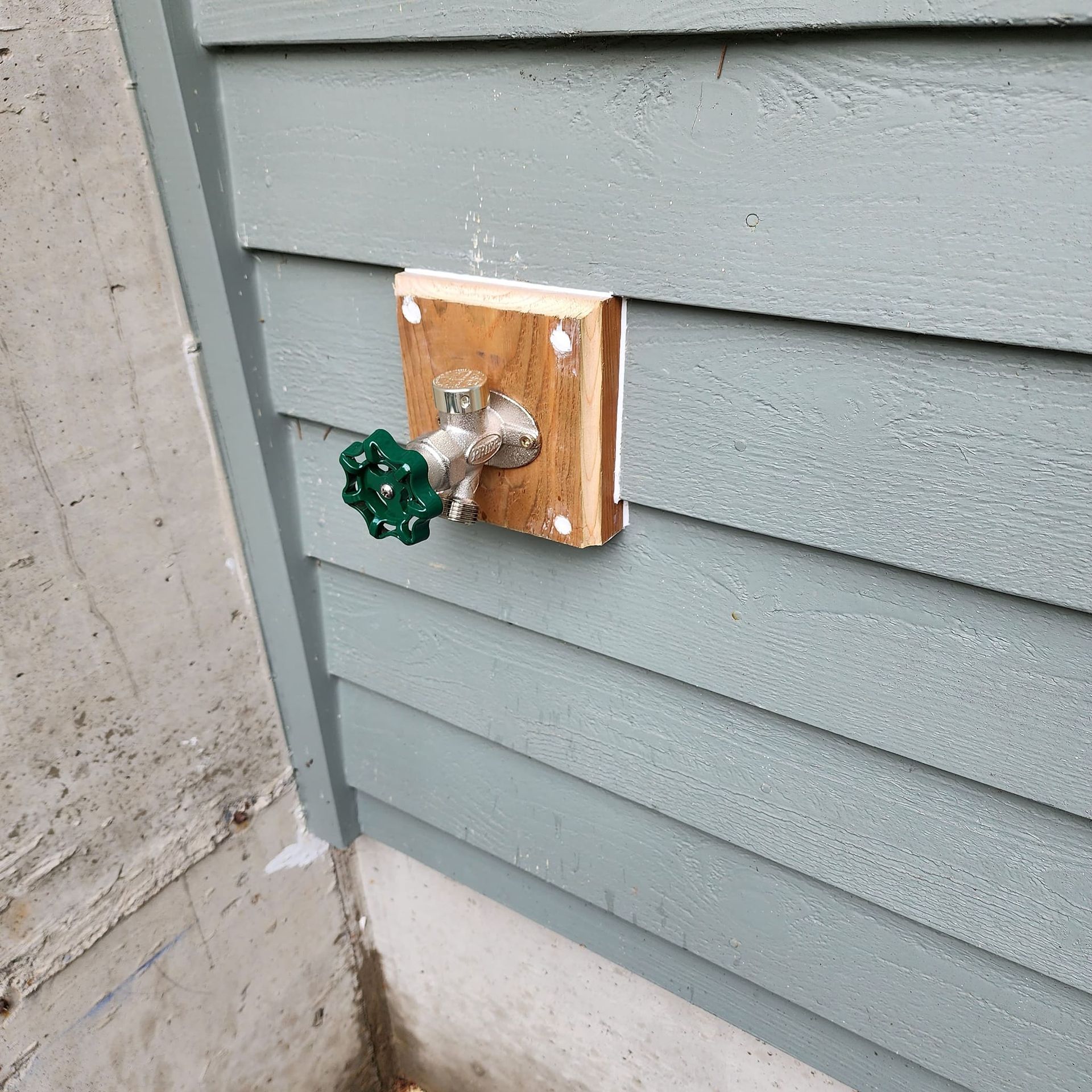 A green-handled outdoor water faucet mounted on a wooden block on a gray clapboard wall.