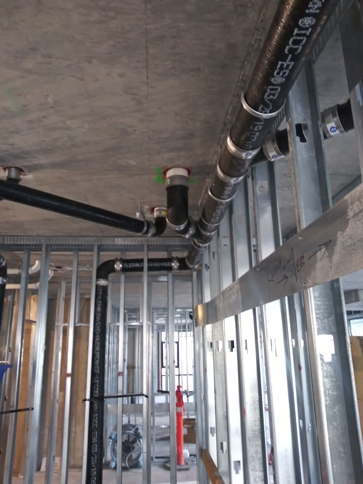 Steel-framed walls with black plumbing pipes installed against a concrete ceiling in a construction setting.