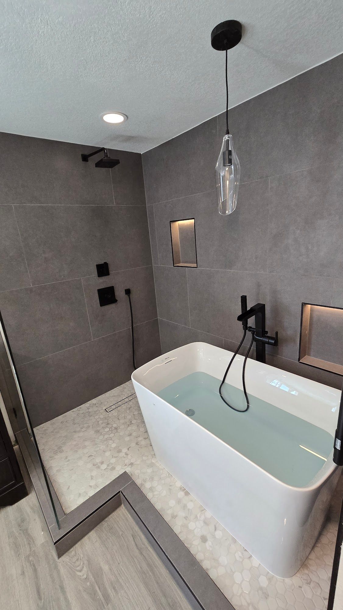 Modern bathroom with gray tile shower, white soaking tub, and black fixtures.