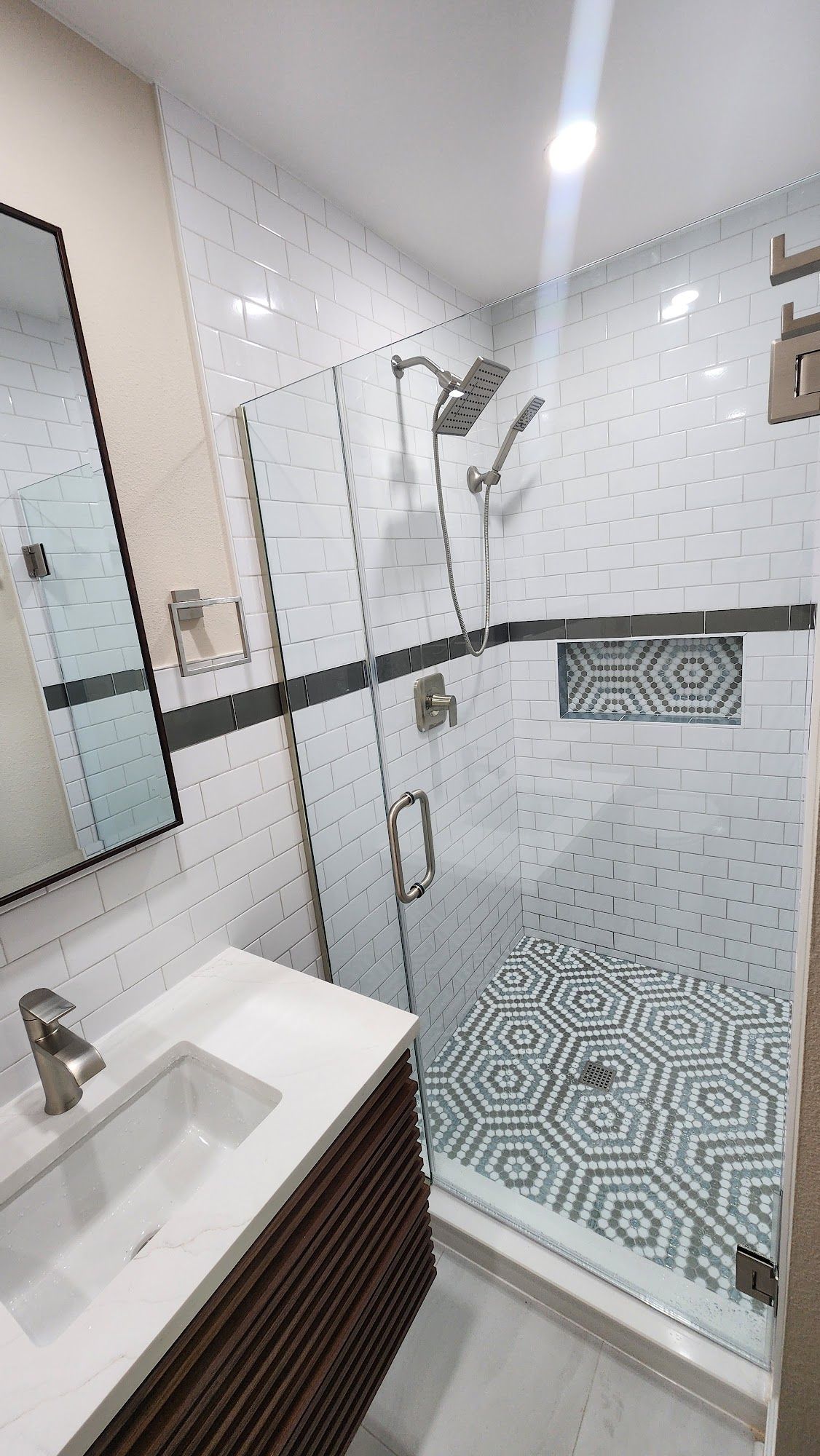 Modern bathroom with glass shower, white patterned tile, dark accent stripe, and a patterned shower floor.