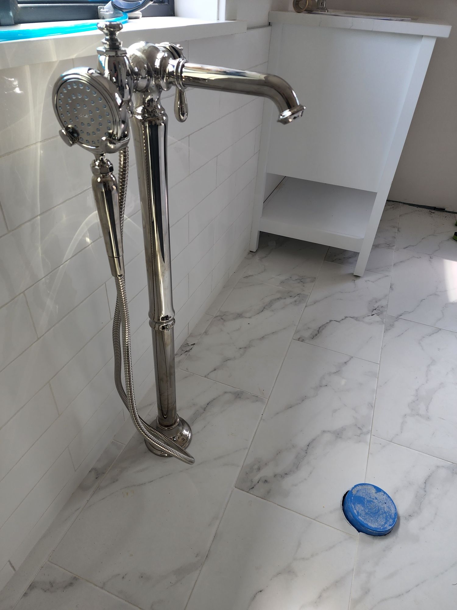 Silver freestanding bathtub faucet on white marble tile floor. White tiled wall and cabinet in background.