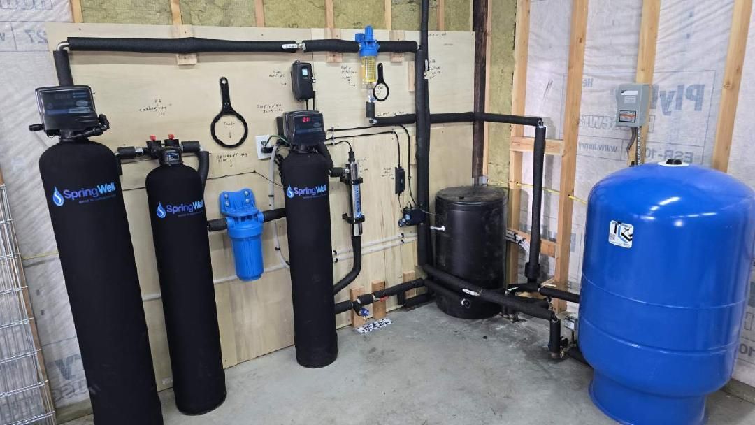 Water filtration system with black tanks, blue filter, pipes, and blue pressure tank in a utility room.