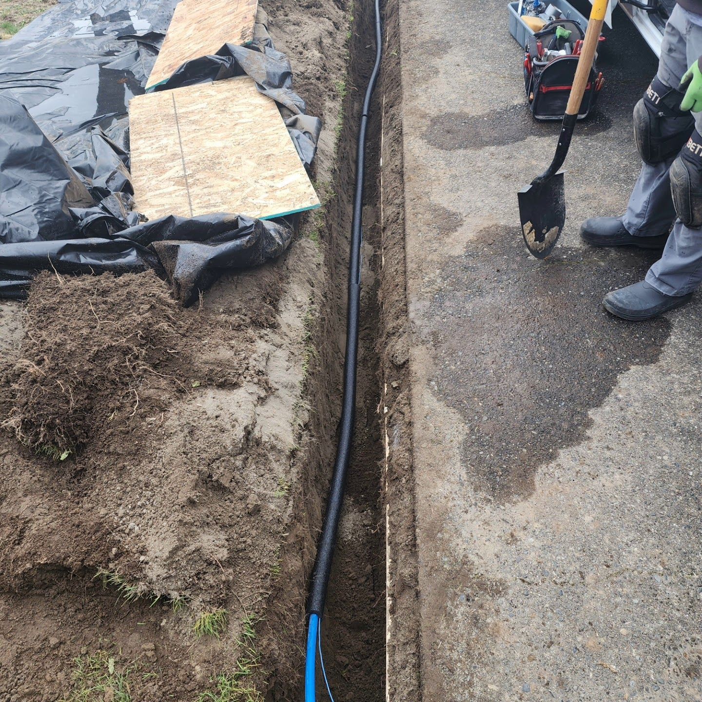 A black conduit in a trench next to concrete. A person holds a shovel.