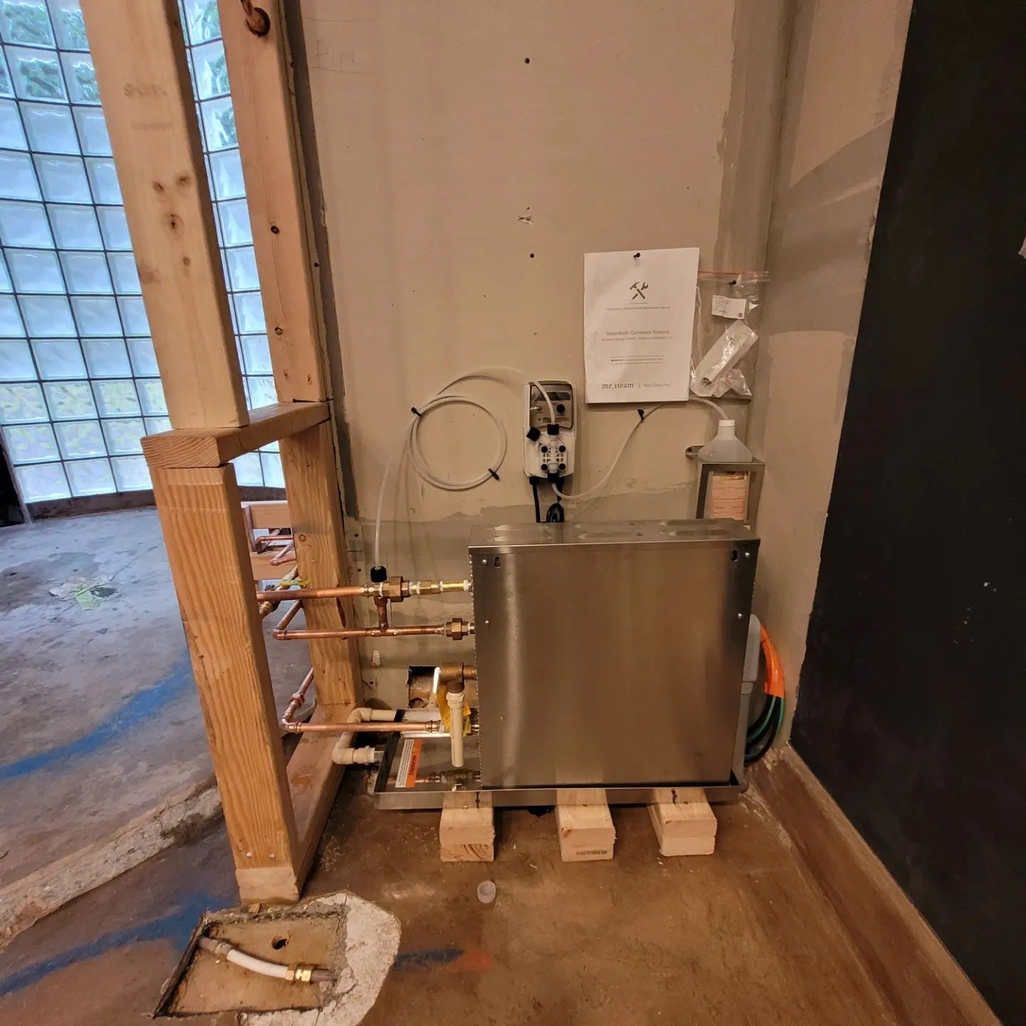 A stainless steel water heater sits on wooden blocks near copper pipes and a partially built wall in a room.