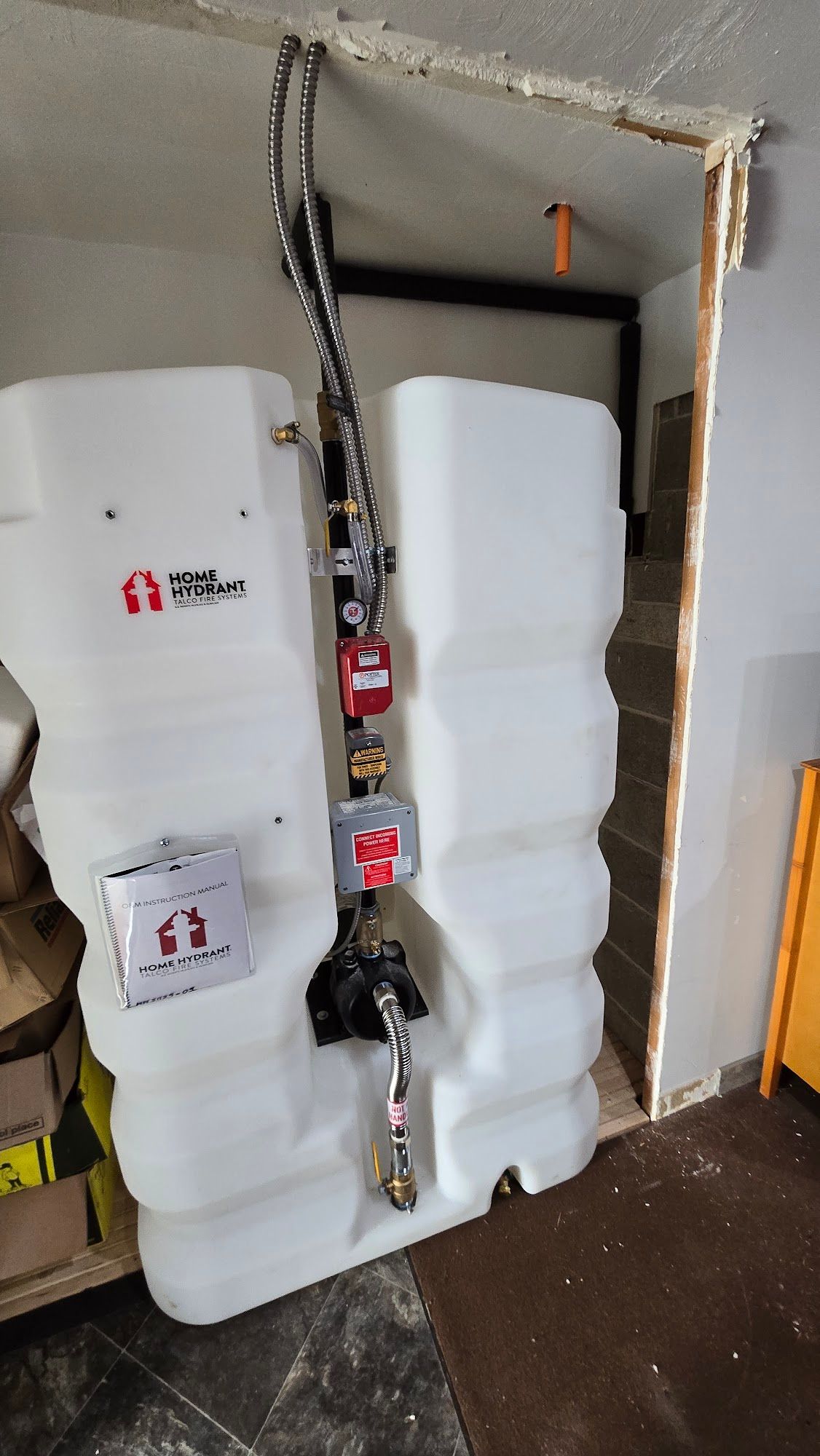 White tank with attached black and silver pipes and valves against a wall.