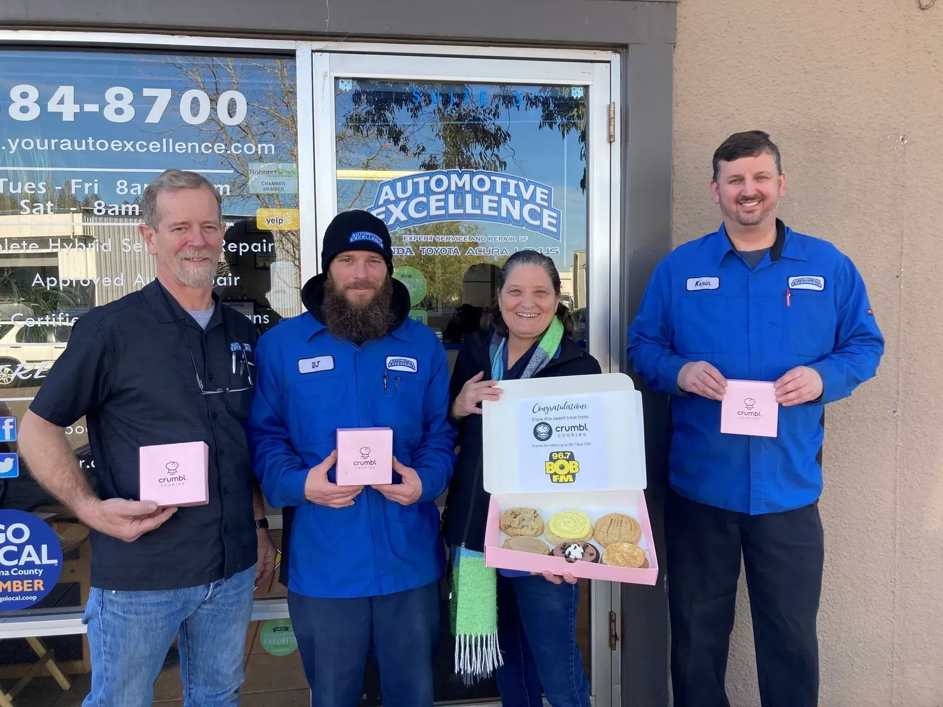 Four people pose outside a shop; two hold donuts, one holds a cookie box. 