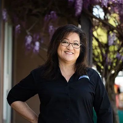 Woman in glasses smiles, standing outdoors near purple flowers, wearing a black shirt. | Automotive Excellence